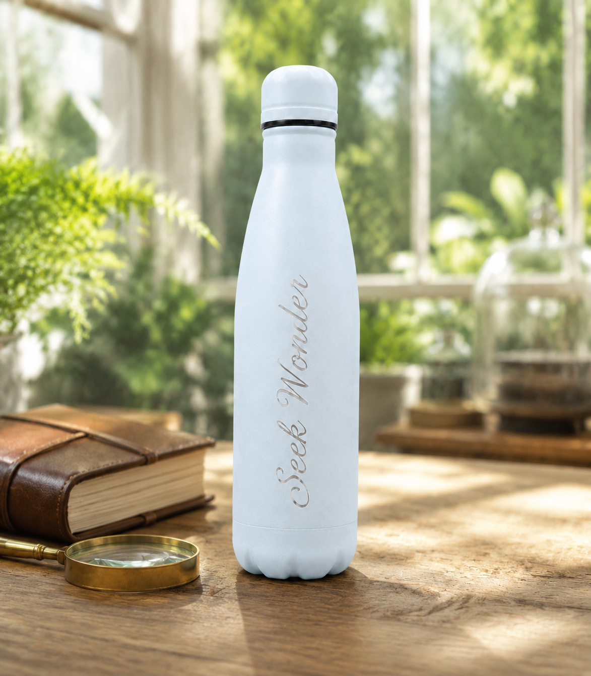 Sky Blue personalised stainless steel water bottle engraved “Seek Wonder” in a sunlit greenhouse with plants, a journal and magnifying glass