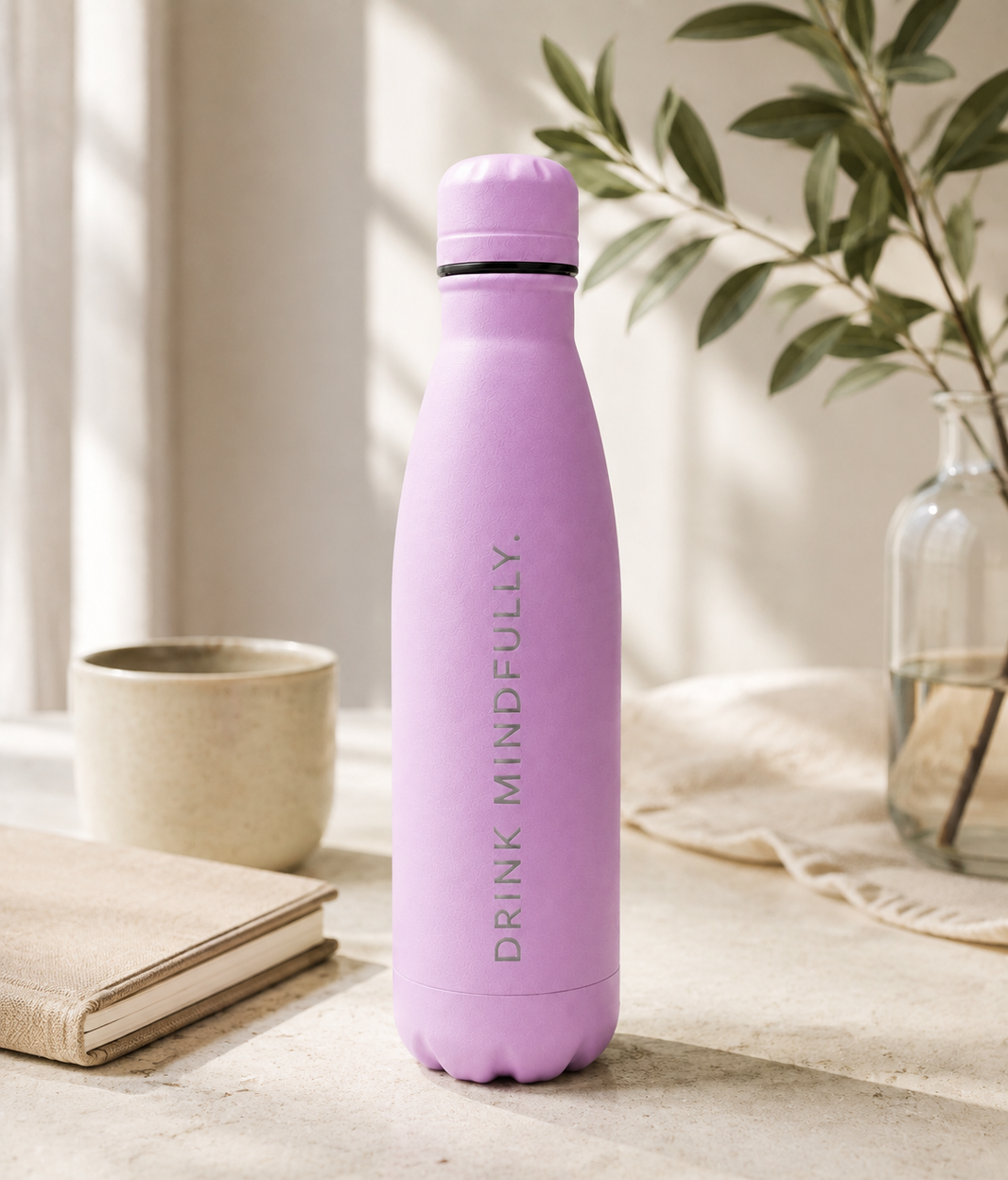 Light purple personalised stainless steel cola-shaped water bottle engraved “Drink Mindfully” on a sunlit home table with a cup, books and leafy vase.
