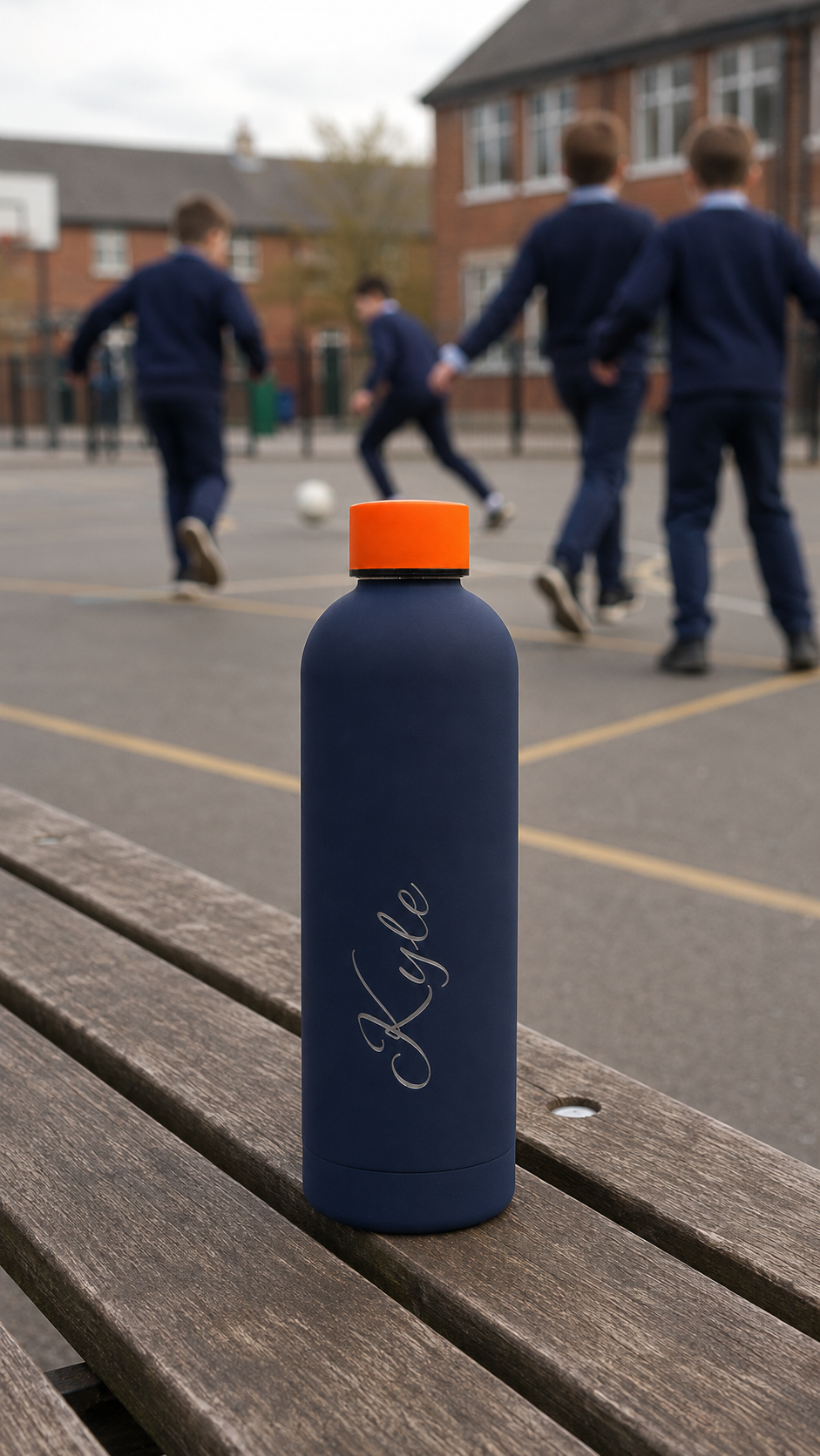 Navy blue personalised stainless steel water bottle with orange lid on a school bench beside children playing football.