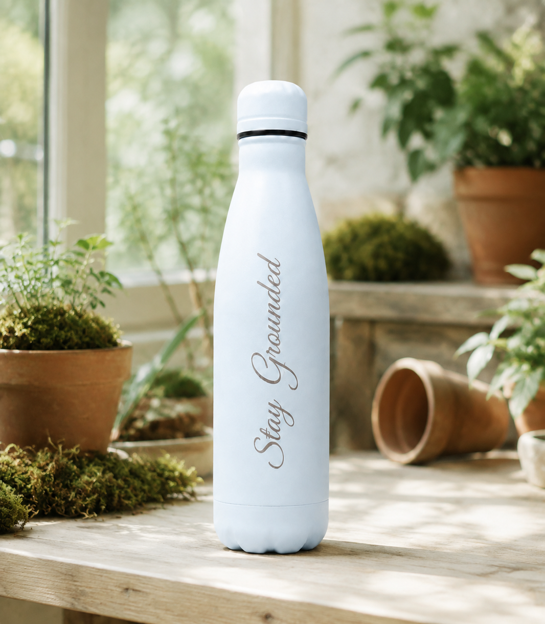 Light Blue personalised stainless steel water bottle engraved “Stay Grounded” on a sunlit indoor garden shelf with potted plants